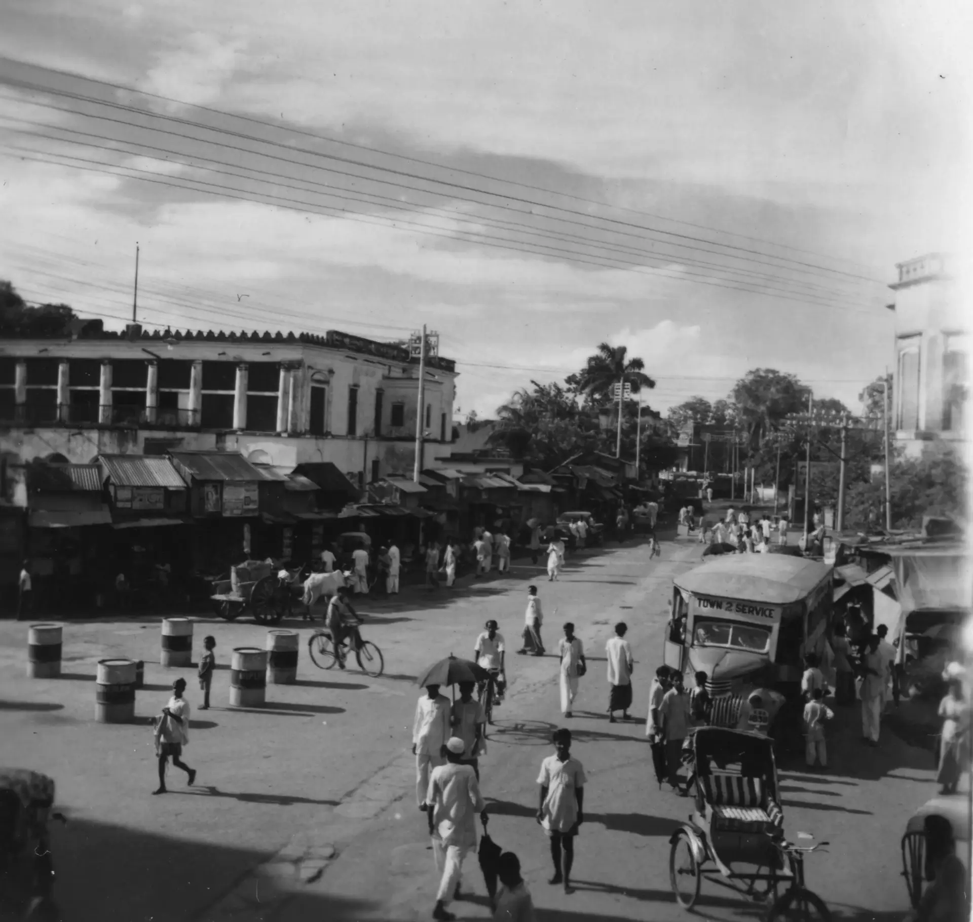 Pedestrians, a cyclist and rickshaws share a Dhaka street with a bus, beneath shopfronts and overhead wires. Photographed in 1952.