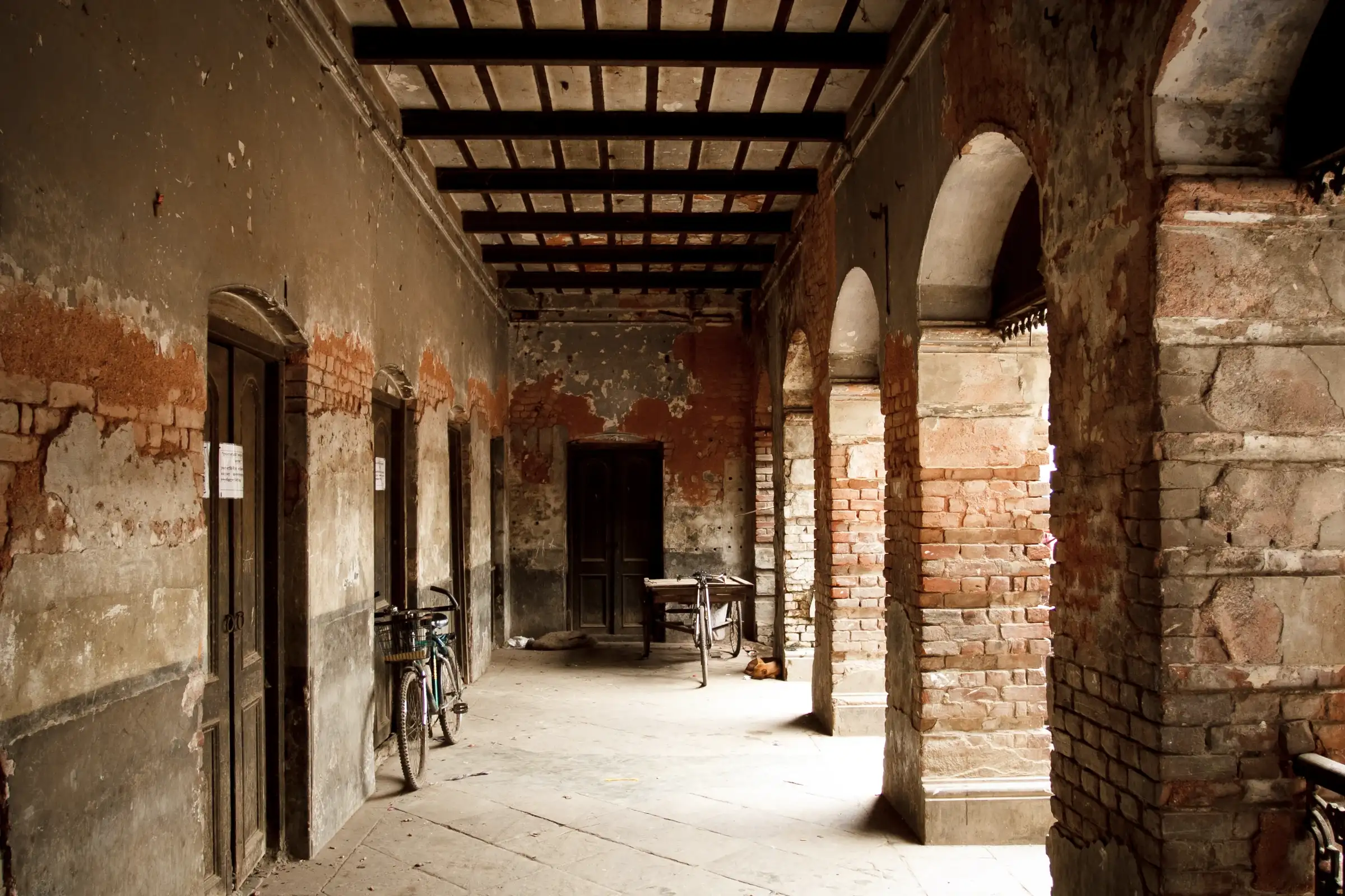 Side-light falls through brick arches onto a passageway at Ruplal House. Wooden doors, two bicycles and a small table stand along the worn plaster walls.